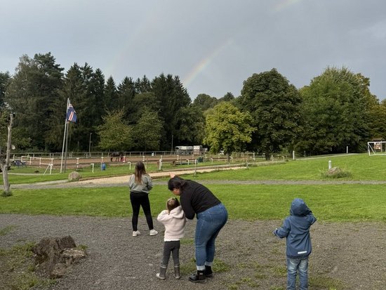 Eine Gruppe von vier Personen, darunter Erwachsene und Kinder, steht auf einem Kiesweg in einem Park. Sie blicken gemeinsam zu einem schwachen Regenbogen, der sich über den bewölkten Himmel und die Baumreihe im Hintergrund spannt.