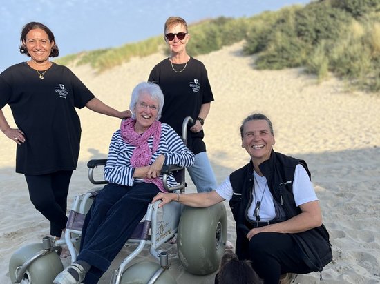 Eine lächelnde ältere Frau sitzt in einem speziellen Strandrollstuhl auf dem Sand. Sie wird von zwei stehenden Frauen und einem knienden Mann umgeben. Im Vordergrund liegt ein Hund mit orangem Geschirr. Im Hintergrund sind Sanddünen und ein blauer Himmel zu sehen.
